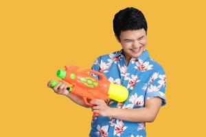 sonriente hermoso asiático hombre jugando con agua pistola aislado en estudio amarillo fondo, Songkran festival. foto