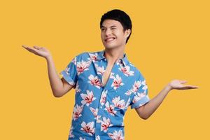 Smiling young handsome Asian man  presenting with open palms gesture on yellow background in studio. He in blue floral shirt for Songkran festival in Thailand. photo