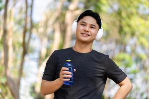 masculino corredor Bebiendo agua después trotar con inalámbrico auriculares en parque foto