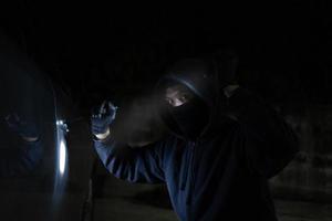 joven ladrón en negro es robando un coche a noche. él lleva un Linterna y un destornillador para coche robo. foto