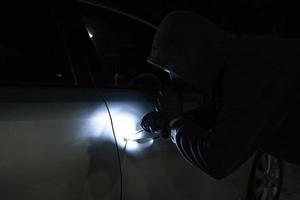 Asian man thief in black mask trying to open car using custom tool and flashlight. Car robber. photo