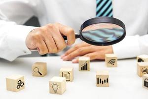 Close up hand of businessperson looking at wooden blocks with various icon symbols through magnifying glass. Concept of business strategy, goal to increase and action plan. photo