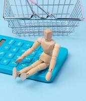 A wooden puppet and a blue calculator, behind a miniature shopping basket photo