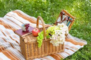 Wicker picnic basket with food on a striped plaid on the grass in the park. Summer picnic concept photo