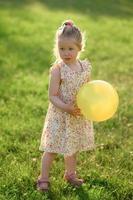 Little girl 3 years old in a summer dress in a clearing with an yellow ball in her hands. The girl is not happy photo