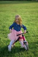 un contento hermosa pequeño niña carreras rápido y paseos su primero bicicleta sin pedales en el césped en un calentar verano día. linda bebé 3 años viejo. lado vista. aprendizaje a mantener balance. foto