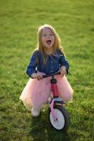 un contento hermosa pequeño niña carreras rápido y paseos su primero bicicleta sin pedales en el césped en un calentar verano día. linda bebé 3 años viejo. lado vista. aprendizaje a mantener balance. foto