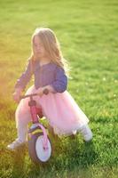 A happy beautiful little girl runs fast and rides her first bicycle without pedals on the grass on a warm summer day. Cute baby 3 years old. Side view. Learning to keep balance. photo