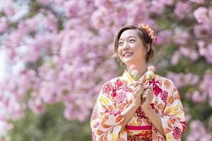 japonés mujer en tradicional kimono vestir participación dulce hanami dango postre mientras caminando en el parque a Cereza florecer árbol durante primavera sakura festival con Copiar espacio foto