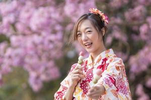 Japanese woman in traditional kimono dress holding sweet hanami dango dessert while walking in the park at cherry blossom tree during spring sakura festival with copy space