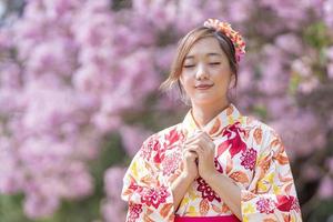 Japanese woman in traditional kimono dress is making a new year wish for good fortune while walking in the park at cherry blossom tree during spring sakura festival with copy space