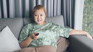 Asian woman smile and happy sit in sofa and holding remote control watching television with relaxing action. Hobby and free time life style concept.