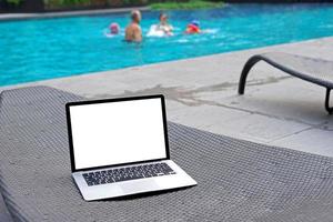 Laptop with blank screen for creative design on sunbed nearby swimming pool background. Computer notebook with monitor clipping path for present landing page design. Laptop computer mock up template photo