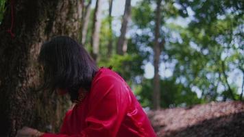 Asian woman in a red shirt standing on a tree trunk in the forest and sweeping a lot of leaf with a broom video