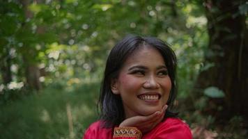 portret van een mooi Aziatisch vrouw in een rood jasje staand in de Woud en glimlachen Bij natuur video