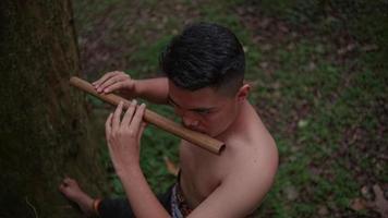 an Asian boy is playing a bamboo flute very seriously in a forest early video