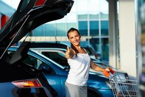 Woman standing with shopping cart and thumb up near the car on the parking photo