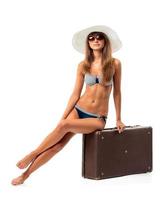 Full length portrait of a beautiful young woman posing in a bikini sitting on a suitcase on a white photo