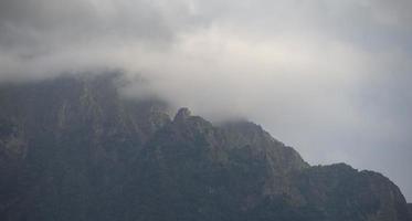 Dark atmospheric surreal landscape with a dark rocky mountain peak in low clouds in a gray cloudy sky. A gray low cloud on a high peak. High black rock in low clouds. Surreal gloomy mountains. photo