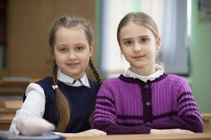 dos colegialas sentar a un escritorio en el salón de clases y Mira a el cámara. foto