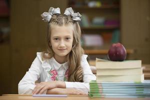 Beautiful schoolgirl with bows at her desk. Girl in the classroom with books and an apple. Secondary school. Back to school. photo