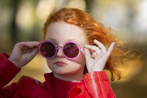 retrato de un elegante Pelirrojo niña en púrpura redondo Gafas de sol. foto