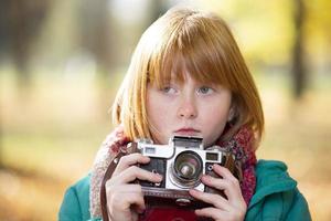 pequeño Pelirrojo niña con un retro cámara en el otoño parque. niño fotógrafo. foto