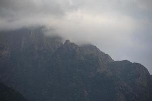 Dark atmospheric surreal landscape with a dark rocky mountain peak in low clouds in a gray cloudy sky. A gray low cloud on a high peak. photo