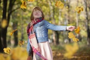 contento Pelirrojo niña dispersa arce hojas en el otoño parque. foto
