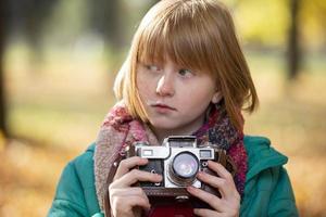 pequeño Pelirrojo niña con un retro cámara en el otoño parque. niño fotógrafo. foto