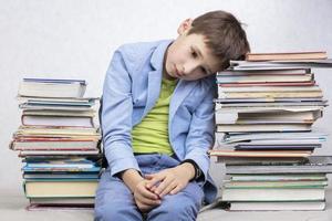 A tired school-age boy sits between stacks of books. Sad child with books. photo