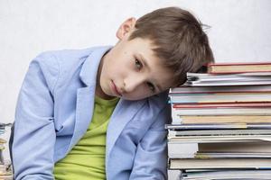 un cansado edad escolar chico se sienta Entre pilas de libros. triste niño con libros. foto