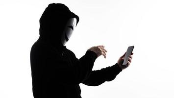 hacker anónimo y mascarilla con smartphone en la mano. hombre con camisa de capucha negra sosteniendo y usando un teléfono móvil con fondo blanco. representan el concepto de piratería informática o robo de datos personales foto