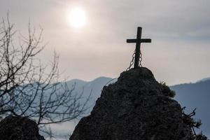 Wooden cross with chain on rock under cloudy sky and bright sun photo