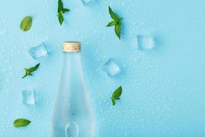 A bottle of ice water, ice cubes, drops and mint leaves on a blue background. photo