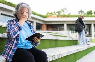 maduro adulto estudiante sentado en frente apagado Universidad edificio y leyendo colegio libros después asistiendo un Universidad clase, adulto educación aprendizaje estudiando contento asiático mayor retirado actividad foto