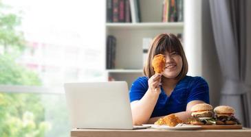 Hungry overweight woman holding Fried Chicken, hamburger on a wooden plate and Pizza on table, During work from home, gain weight problem. Concept of binge eating disorder photo