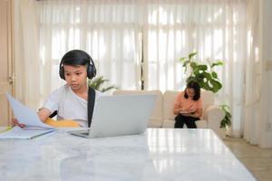 Asian boy playing guitar and watching online course on laptop while practicing for learning music or musical instrument online at home. Boy students study online with video call teachers play guitar. photo