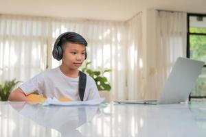 Asian boy playing guitar and watching online course on laptop while practicing for learning music or musical instrument online at home. Boy students study online with video call teachers play guitar. photo