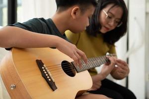 Asian boy playing guitar with mom in the living room for teaching him son play guitar, feel appreciated and encouraged. Concept of a happy family, learning and fun lifestyle, love family ties photo