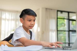 Asian boy playing guitar and watching online course on laptop while practicing for learning music or musical instrument online at home. Boy students study online with video call teachers play guitar. photo