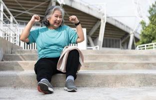 Happy and smile elderly asian woman sitting on stairs for rest after workout, jogging on morning, senior exercise outdoor for good healthy. Concept of healthcare and active lifestyle for healthy