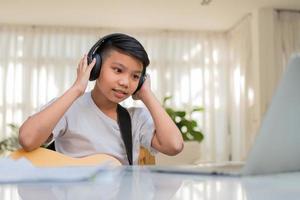 Asian boy playing guitar and watching online course on laptop while practicing for learning music or musical instrument online at home. Boy students study online with video call teachers play guitar. photo