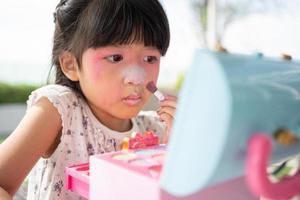 adorable pequeño niño asiático niña pinturas su boca con rosado niños cabezas y mira en el espejo. un niño obras de teatro a hogar en un juguete belleza salón. incrementar aprendizaje desarrollo para preescolares foto