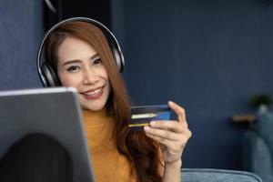 retrato de joven mujer vestir auriculares y participación tableta y bancario crédito tarjeta, transferir dinero en línea, compras bienes en Internet almacenar, adquisitivo servicios, satisfecho con seguro pagos foto