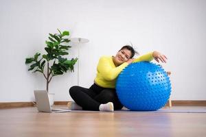 Portrait fat woman rest after exercise with fitness ball in home gym. Overweight woman sitting on a pilates ball and Stretching her muscles before exercising. Health care and weight loss concept. photo