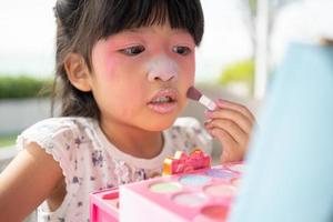 adorable pequeño niño asiático niña pinturas su boca con rosado niños cabezas y mira en el espejo. un niño obras de teatro a hogar en un juguete belleza salón. incrementar aprendizaje desarrollo para preescolares foto