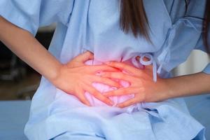 Unhappy Asian patient woman sitting on the hospital bed and holding on stomach suffering. Abdominal pain that comes from menstruation, diarrhea, or indigestion. Sickness and healthcare concept photo