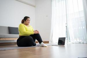 Asian overweight woman doing stretching exercise at home on fitness mat. Home activity training, online fitness class. Stretching training workout on yoga mat at home for good health and body shape. photo