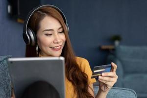 retrato de joven mujer vestir auriculares y participación tableta y bancario crédito tarjeta, transferir dinero en línea, compras bienes en Internet almacenar, adquisitivo servicios, satisfecho con seguro pagos foto